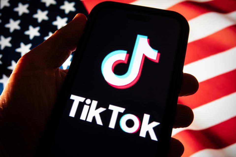 A user holding a phone with TikTok logo on the screen and American flag in the background.